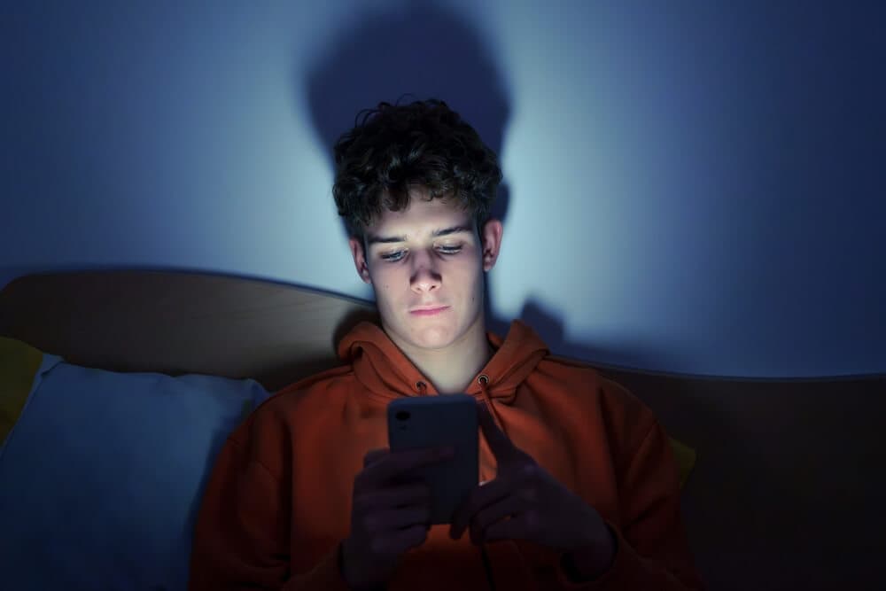 Teenager lying in bed scrolling on smartphone in a dark room, screen light illuminating their face